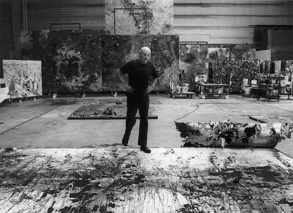 A black and white photograph of the artist Anselm Kiefer. It show a white male standing in the centre of the photograph in an artists studio. There are artworks on the floor and around the walls. He is standing with one foot on an artwork on the floor. He is wearing black trousers shoes and a black t-shirt. His hands are on his hips. 