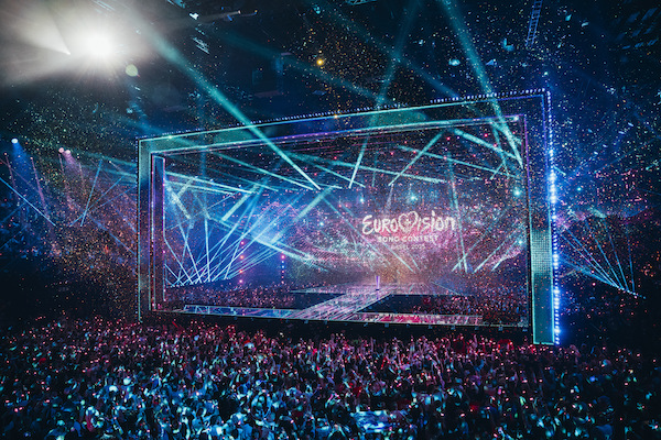 Photograph of an indoor concert. The fans standing below the stage hold up multi-coloured lights. Bright lights in green project out into the auditorium. The words Eurovision Song Contest are on the screen at the back of the stage. 