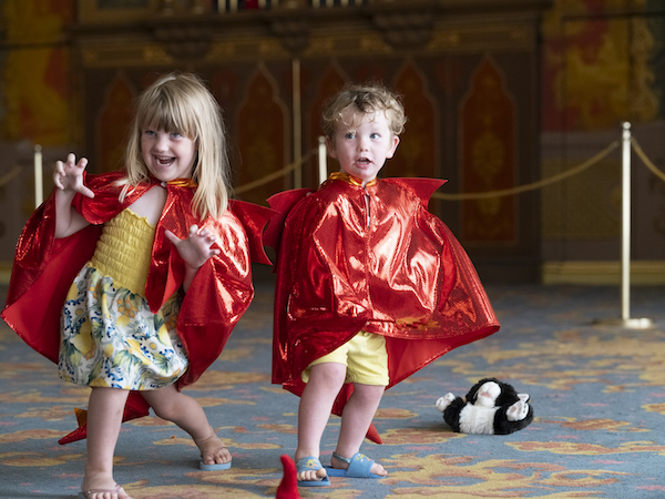 Two young children in red dragon cloaks, under the cloaks they wear yellow summer clothes and blue summer sandals. There is a blonde girl on the left making a animal stance with fingers curled as she growls. The little boy is younger and looks off to the left. There is a lonely black and white teddy left on the blue and gold carpet. 