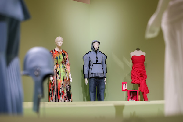 Photograph of a costume display. Three outfits are seen against a light green wall on mannequins. The far right is a red strapless dress, the middle has a grey hoodie and jeans and the left a multicoloured red and green robe. 