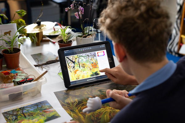 A young person sits at a desk, we see him from behind. they have light brown curly hair. On the desk is an ipad with an image of Rousseau's painting 'Surprised. On the desk are copies of the Rosseau art work and a few plants in small brown pots. 