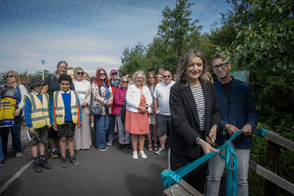A photograph of a man and a woman cutting a blue ribbon outside. Behind the two people stand members of the public and school children who are smiling. There is blue sky and green bushes behind the crowd. 