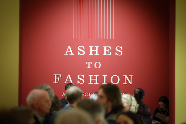 Photograph of the opening of Ashes to Fashion. White wall text on a red background shows the words 'Ashes to Fashion'. Underneath the text heads of visitors can be seen moving to and fro.  