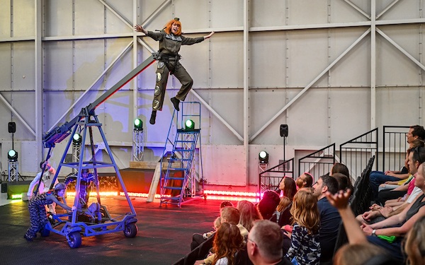 An actress dressed in a khaki green jumpsuit and orange hair is suspended in the air by a metal frame she has her arms outstretched like she is flying. To the right is tiered seating with families watching the show.