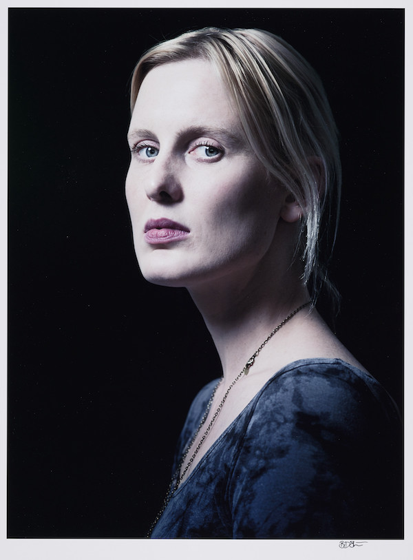 A colour photograph of a white woman showing her head and shoulders. She body is facing the left and she is looking towards the camera, she has fine white blonde hair in a low ponytail and pale skin. She wears a blue top and a chain necklace. The woman is the author Amy Liptrot who wrote The Outrun.