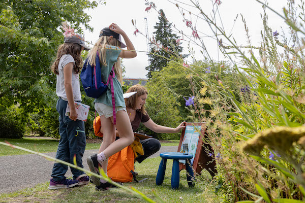 In a garden a family group are looking at an information board. A woman is crouching down reading the information two children stand to her left the both wear caps and t-shirts and trainers they are carrying paper trails. There is greenery and flowers behind them and to the left of the image. 