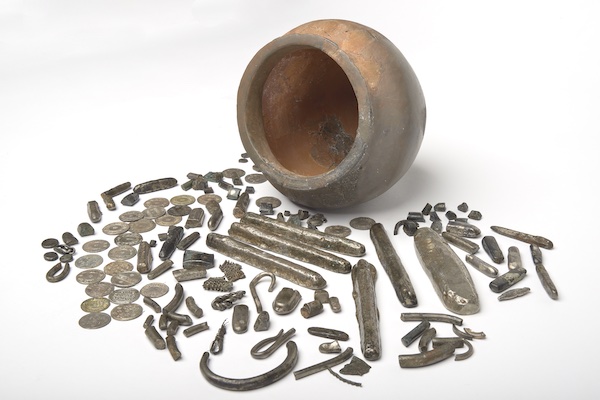 Photograph of an empty terracotta pot lying on its side. In front of the pot are an assortment of metal objects including coins, ingots and fragments of jewellery.. 