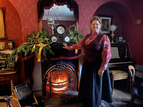 A woman stands in a old fashioned living room in front of a roaring fire. She wears a long dark skirt, maroon waistcoat and patterned blouse. On top of the mantel there are seasonal green branches, pine cones and yellow ribbon.