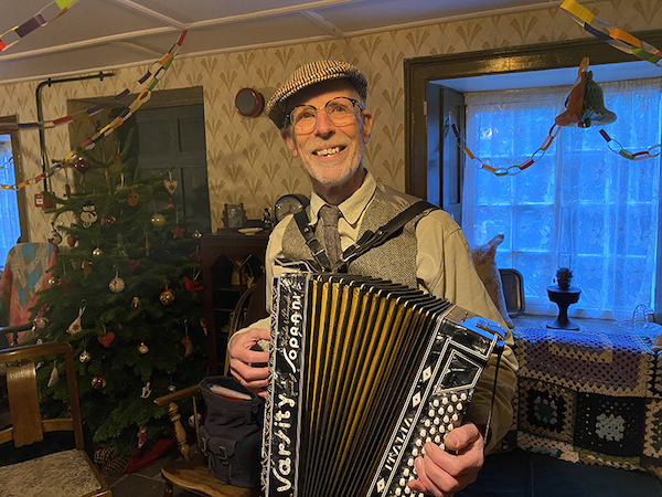 A man stands in an old fashioned front room, decorated with paper chains and a traditional Christmas Tree, he wears a brown flat cap and waistcoat and glasses. In his hands is an accordion.