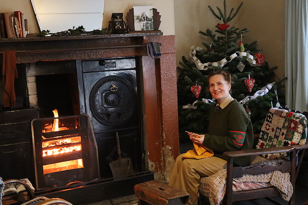 A woman sits in a low arm chair in a house decorated in 1940s style. She wears beige and green army clothes and knits in yellow wool. She sits in front of a black range with a warming fire. Behind her there is a traditional Christmas Tree decorated with paper chains. 