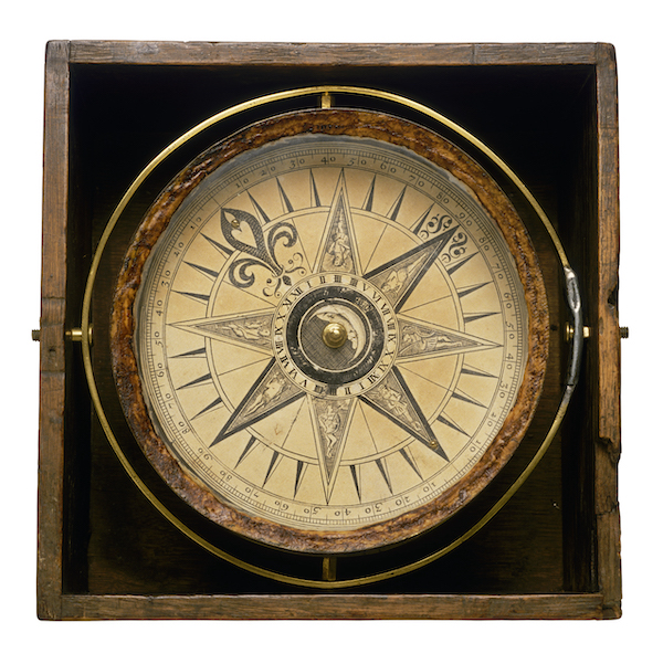 A photograph of a compass set in a wooden box. The compass has a circular metal surround, white and black split colour triangles mark regular intervals on the outer circle. In the centre is an 8 pointed star with roman numerals visible on an inner circle.