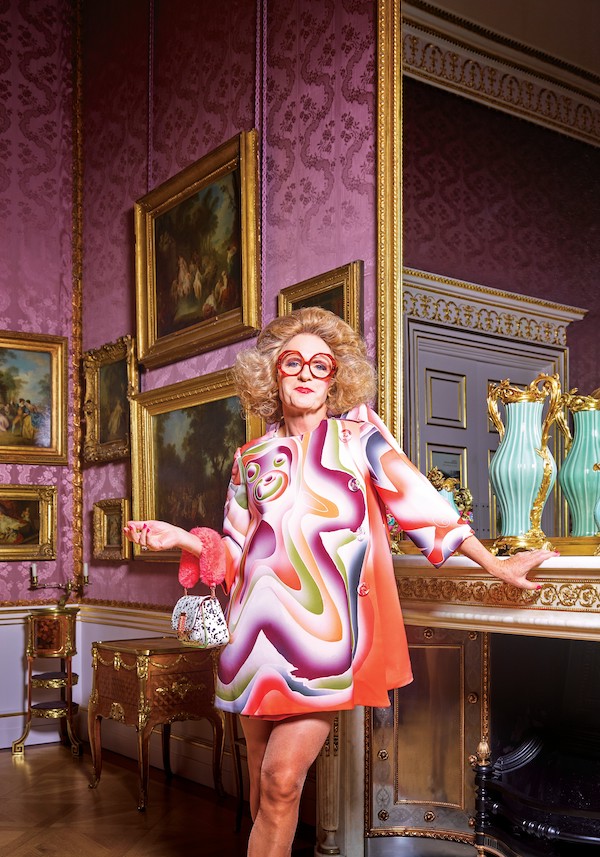 Photograph of the artist Grayson Perry, dressed as his alter ego. It shows a woman standing at the Wallace Collection facing the camera their left hand is resting on the fire place, their right hand is bent at the elbow and holds a small handbag. They have big blond hair in a bouffant style, large red framed glasses and a short 60s style psychedelic dress in a wavy pattern of orange purple and green. 