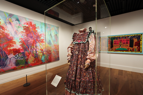 Photograph of the Grayson Perry exhibition at the Wallace Collection. The photograph is inside the gallery space it shows a glass case featuring one of the artist Grayson Perry's dresses that he wears as his alter ego. On the walls behind the case there are Grayson Perry artworks. They are bightly coloured set agains a white walls and a wooden floor. 