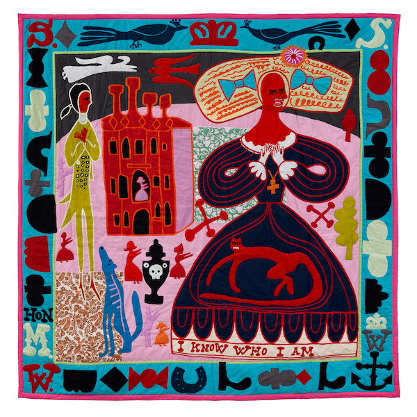 A fabric artwork showing embroidery applique that shows an illustrated version of the artist Grayson Perry as his alter ego. The artist wears a dark of dark blue with red lines and an image of a red animal on the skirt. The work in bordered in a light blue with symbols including anchors crown and peacocks. On the bottom of the artist's dress is a white border saying 'I know who I am'. There is also a tall building in the background that looks like a castle and a man dressed as a footman or servant with old fashioned dress. 