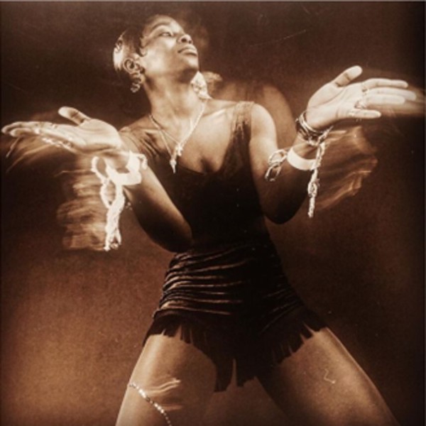 A sepia-toned photograph of a young woman dancing against a dark background. She wears shorts and a crop top with tassle fringing, with lots of jewellery on her ears, neck and wrists. Her hands are pointed outwards as she dances, looking to the side. The image has a slight blur due to the long exposure time.