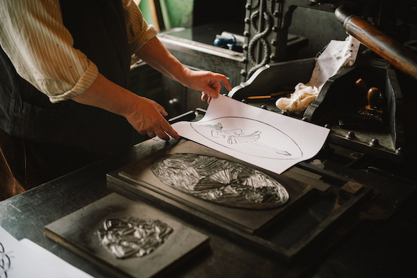 A man stands next to a table from the photo we only see the neck down. The man is printing using a wood block print. In his hands he is lifting off a piece of paper with a black outlined image. He wears a white shirt rolled up at the sleeves and a waistcoat.