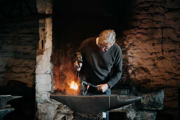 A man stands over an anvil in his hands are large metal tools a hammer in his right hand and a pair of pincers in his left. Behind him there is a small fire, he ears and dark wooly jumper, he has grey hair and glasses. He stand in a building with rough stone walls.  