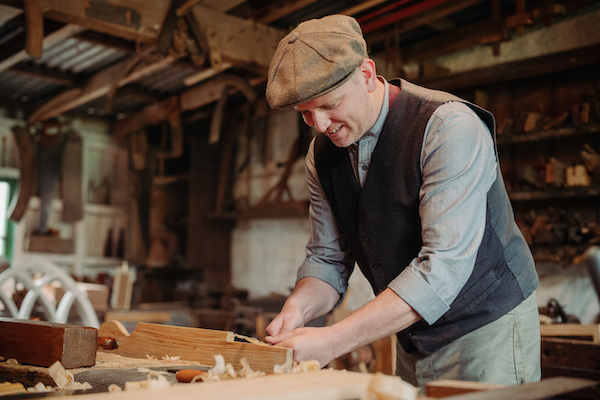 A man stands over a bench, he is using his hands and working with light coloured wood on the bench. There are wood shavings around his work. He wears a waistcoat, a shirt with his sleeves rolled up and a flat cap. He is smiling. Behind him are tools and it looks like a wood working workshop. 