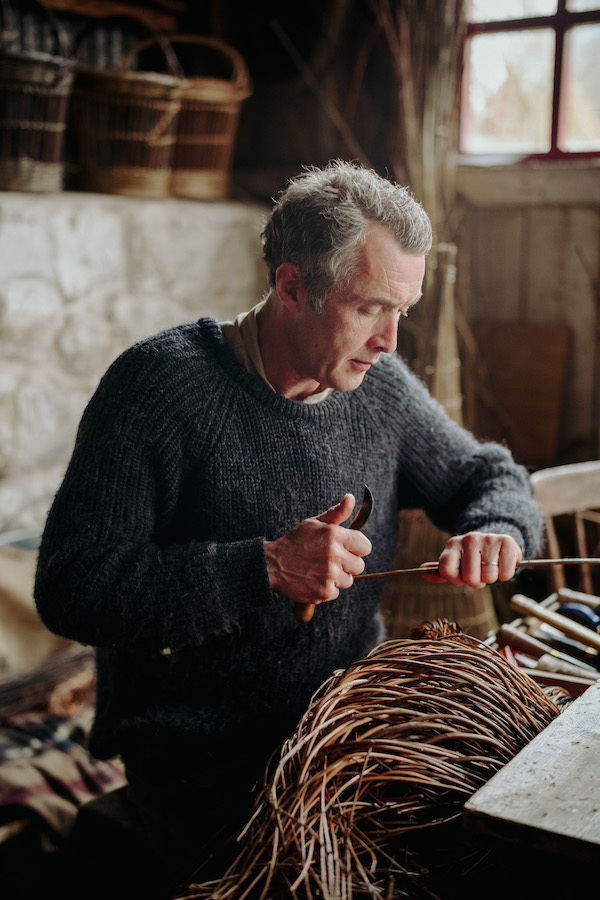 A man stands in front of willow branches, he holds a branch in his left hand and in his right he has a small curved knife. The man is wearing a woolly jumper and works in a cottage.  