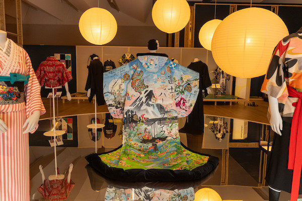 Photograph of the Kimono exhibition at V&A Dundee in the centre is displayed a blue kimono with a mountain scene, there are a number of mannequins displaying kimonos in the background the space is lit by large paper ball lanterns. 