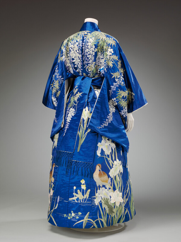 A photograph of a blue kimono from the back. It has scenes of nature including flowering wysteria across the shoulders and a wading bird on the bottom. It is very beautiful.