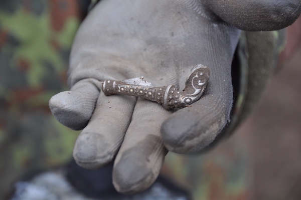 Photograph of a hand wearing a blue glove in the hand is a silver brooch about the width of the hand in size it is silver and ornate.