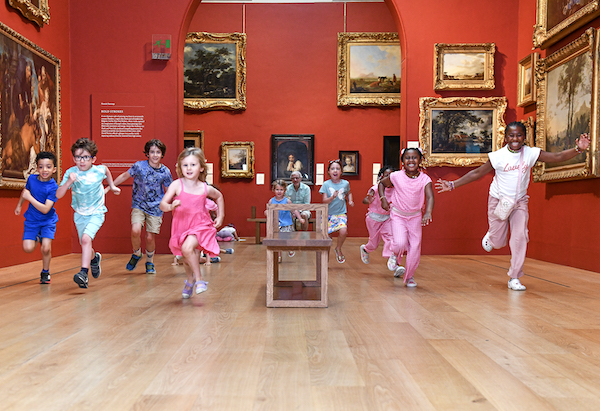 In a red walled are gallery there are paintings with gold frames on the walls. A group of 9 children in summer clothes run through the gallery towards the camera.