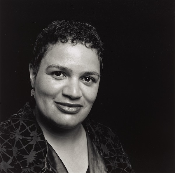 A photograph of a black woman with short close cropped hair. She stares directly at the photographer with a slight smile on her lips. The woman is Jackie Kay the Scottish poet and play write. 