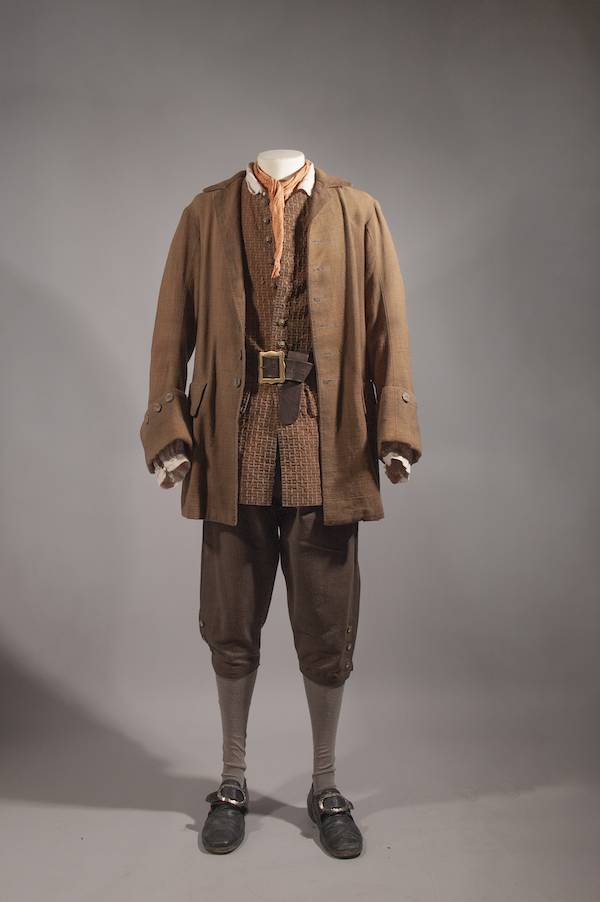 A photograph of a film costume belonging to the character Will Turner, played by Orlando Bloom in the film - Pirates of the Caribbean. It is a brown jacket, waistcoat and breaches with black shoes and an orange necktie.