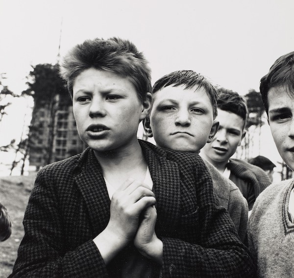 A black and white photo of 4 young lads. The boy in front is shown from the chest up, his hand clasped in front of his chest, he has short bushy hair. He looks off to the left and wears a smart jacket with a t-shirt. The boys behind him crowd round and stair at the photographer. In the background you can see a tower block.  