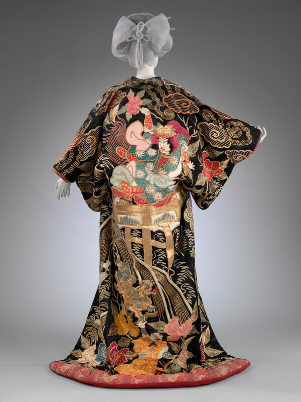 Photograph of the back of an outer-kimono for a woman it is black and gold with a red band across the bottom the motifs relate to a kabuki play and show a figure and stone bridge.