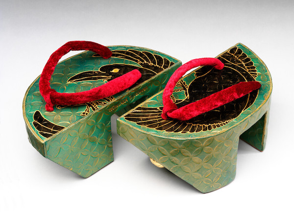 Photograph of a pair of shoes, they are a type of wooden sandal with a green painted base and red fabric thong. A black raven is painted on the surface of the shoe.