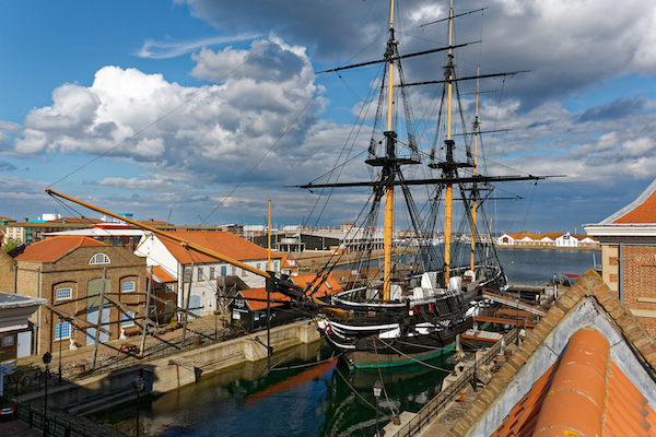 Photograph of a sail ship in dock surrounded by houses. The ship is black and white with a yellow masts. 