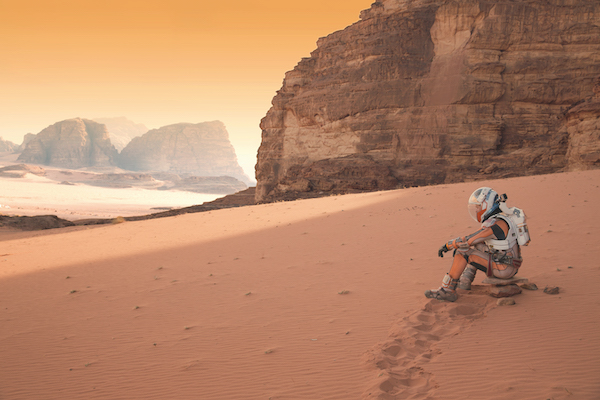 A film still from the Martian. A astronaut sits on red sand. His knees are bent and his arms rest on his knees. The landscape shows red rocky mountains. It is desolate and there are no other people or buildings. 