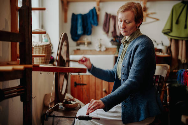 A woman is weaving, she wears a blue cardigan and green shirt, she has short hair. She is weaving thread using a wooden loom. She stand in a rustic room with coloured yarn hanging on the walls. 