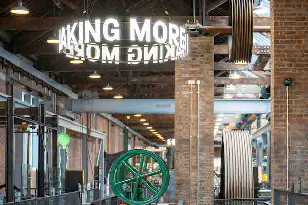 Photograph of Power Hall a large open museum space with a wooden hall and brick pillars. A yellow train is visible to the left and right. Wooden roof beams criss cross the top of the photo and a white circular sign lights up the centre of the photo.