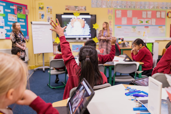 The photograph shows a primary school classroom with bright yellow walls which are covered with signs and posters. Two teachers stand either side of an interactive white board that has an image of Queen Elizabeth I. The children in red jumpers sit at desks, two of the children can be seen looking at ipads, one child has their hand up to answer or ask a question. 