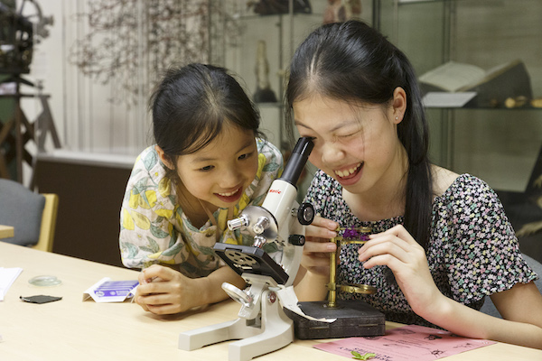 Two children sit at a table. One girl looks into a microscope and another sits excitedly at her side. 