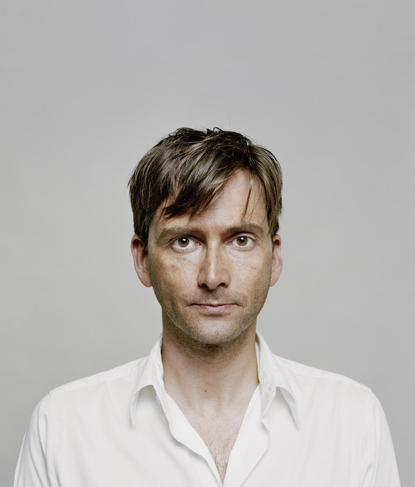 A colour photograph of a middle aged man he stares straight at the camera. He has brown eyes and brown short hair. He wears a white open necked cotton shirt. The man is the actor David Tennant.